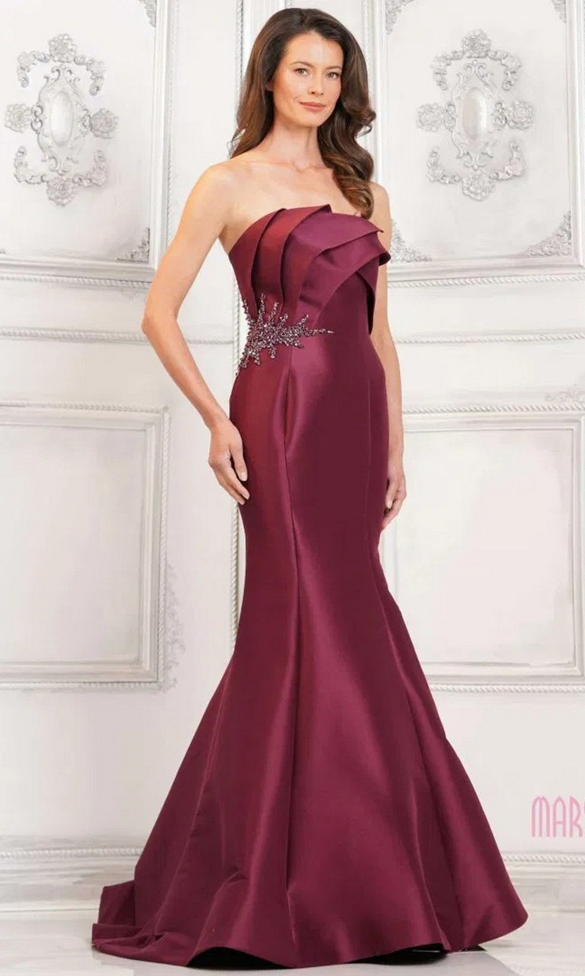 A woman stands in an ornate white room wearing the Marsoni MV1308, a long burgundy formal dress featuring a strapless mermaid silhouette with pleated detailing and beaded applique at the waist.